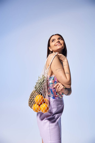 Una giovane donna elegante con i capelli castani in un vibrante abito viola che tiene una borsa di frutta fresca assortita. - Foto, immagini