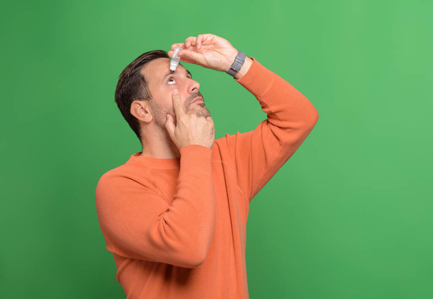 Young man dropping eye lubricant to treat dry eye or allergy while standing on green background - Photo, Image
