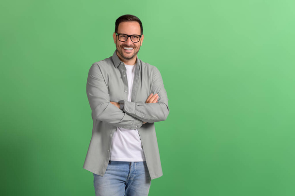 Portrait of smiling handsome entrepreneur in eyeglasses and with arms crossed on green background - Photo, Image