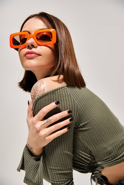 Una giovane donna chic con i capelli castani che indossa occhiali da sole rossi e un top verde in un ambiente da studio. - Foto, immagini