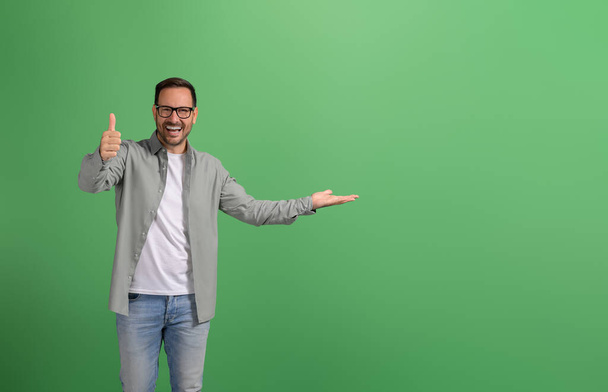 Salesman laughing and demonstrating product on palm and showing thumbs up sign on green background - Photo, Image