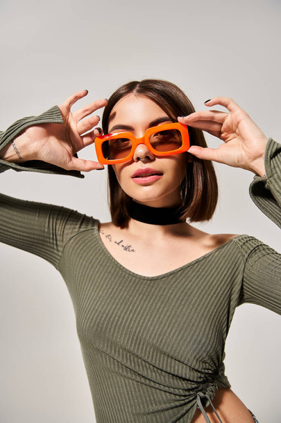 Стильна молода жінка з брюнеткою у зеленій сорочці та помаранчевих сонцезахисних окулярах у студійній обстановці. - Фото, зображення