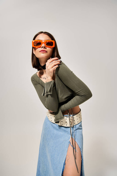 Een jonge vrouw met brunette haar poseert vol vertrouwen voor een foto in een stijlvolle rok en zonnebril. - Foto, afbeelding