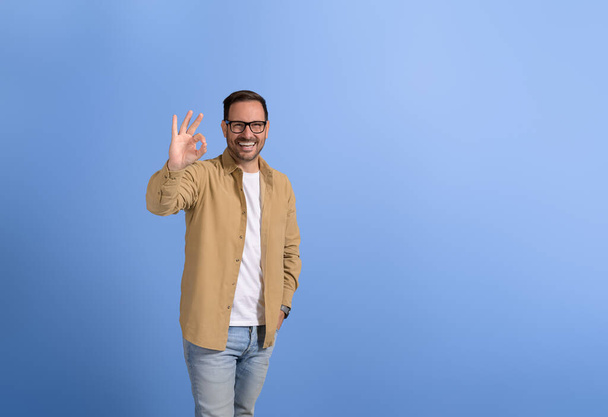 Portrait de beau vendeur avec la main dans la poche souriant et montrant OK signe sur fond bleu - Photo, image