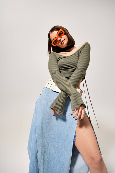 A young woman with brunette hair strikes a stylish pose in a studio while wearing a skirt and sunglasses. - Fotó, kép