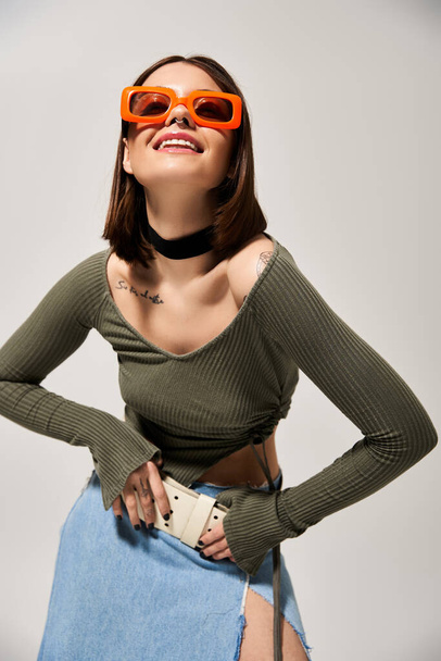 A fashionable young brunette woman confidently wears sunglasses and a stylish skirt in a studio setting. - Photo, Image