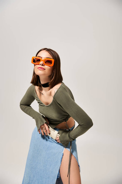 Μια κομψή νεαρή μελαχρινή γυναίκα ποζάρει με αυτοπεποίθηση σε φούστα και γυαλιά ηλίου για μια φωτογραφία. - Φωτογραφία, εικόνα