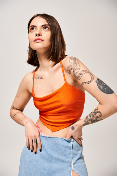 Een jonge vrouw met brunette haar die een zelfverzekerde houding aanneemt, handen op de heupen, in een levendige oranje top in een studio setting. - Foto, afbeelding