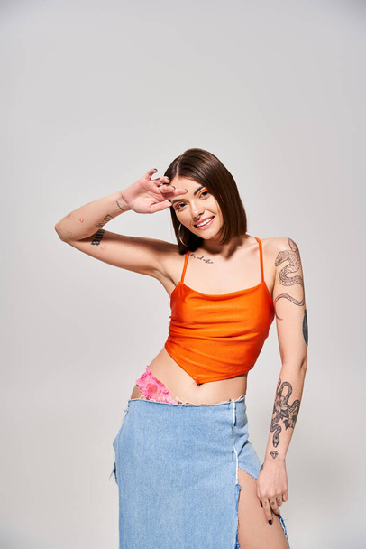 A young woman with brunette hair confidently poses in a studio wearing an orange top and a flowing blue skirt. - Photo, Image