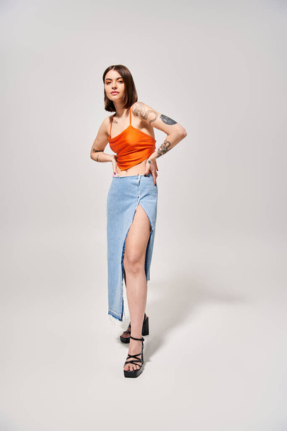 Een stijlvolle jonge vrouw met brunette haar staat sierlijk in een studio met een oranje topje en een vloeiende blauwe rok. - Foto, afbeelding