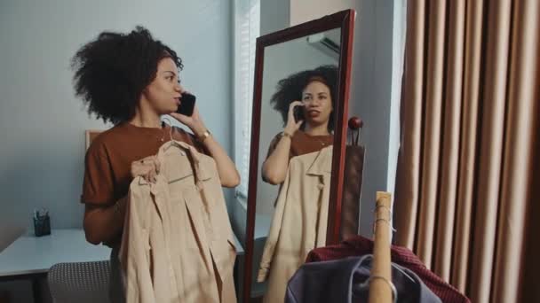 Medium long shot of young curly woman talking on mobile phone while trying on beige coat near mirror at home - Footage, Video