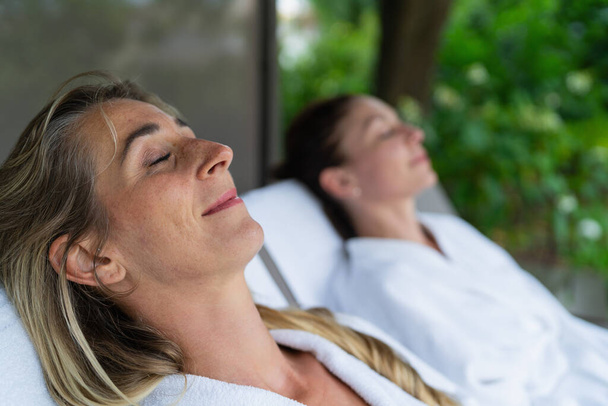Two women relaxing on outdoor loungers, one in focus with eyes closed. Spa wellness hotel concept image - Photo, Image