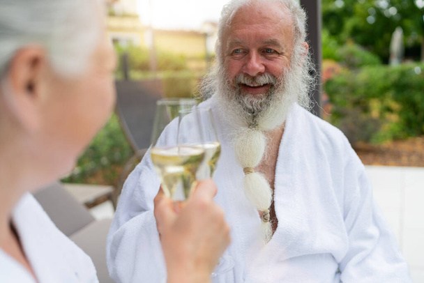 Ηλικιωμένος άντρας με λευκή γενειάδα χαμογελά και κάνει πρόποση με ένα ποτήρι σαμπάνια - Φωτογραφία, εικόνα