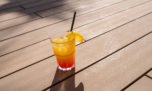 Tropischer Cocktail mit Orangenscheibe auf Holzdeck, Sonnenschein wirft Schatten, erfrischendes Getränk in einem karibischen Inselhotel - Foto, Bild