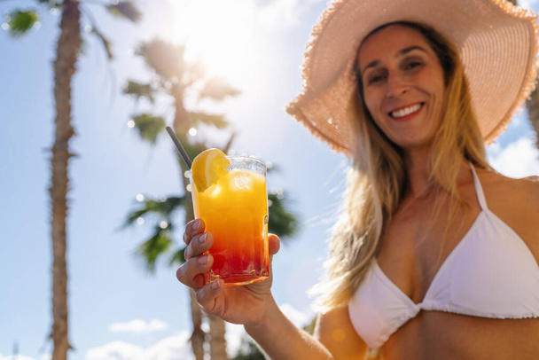 Woman in white bikini and straw hat offering a tropical cocktail, sunny sky with palm trees. Party and summer holidays concept image on a caribbean island. - Photo, Image