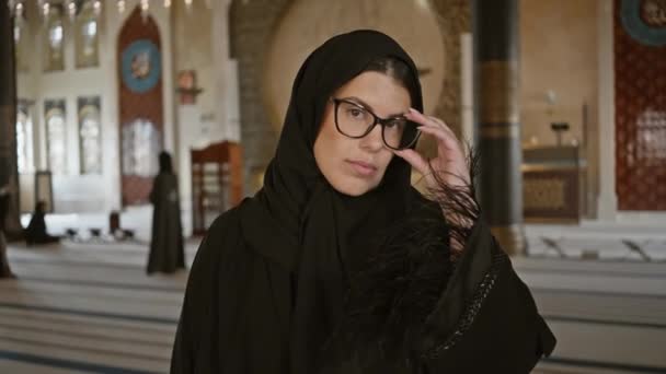 Молодая взрослая женщина с теплой улыбкой, в очках и традиционном хиджабе, позирует в декоративном интерьере мечети Катара, Катар. - Кадры, видео