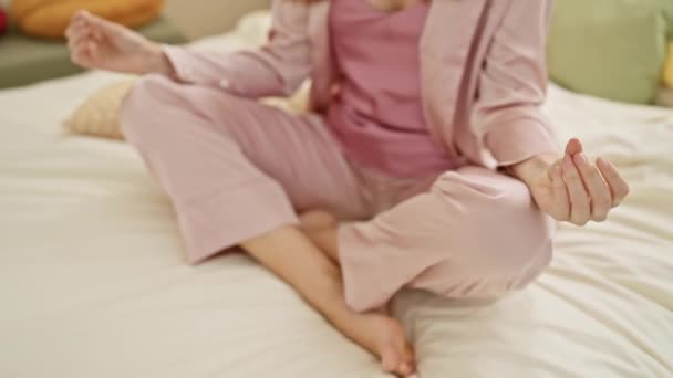 Młoda ruda kobieta w różowej piżamie medytuje na łóżku w jasno oświetlonej sypialni - Materiał filmowy, wideo
