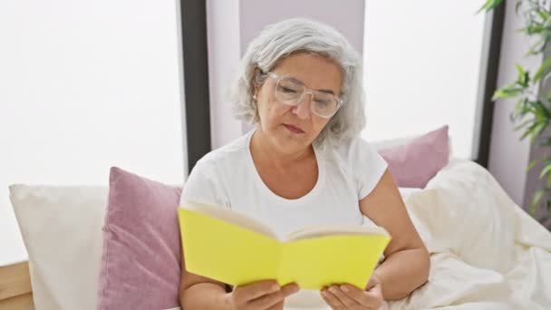 A mature woman with grey hair and glasses relaxing in her bedroom at home while reading a yellow book. - Filmati, video