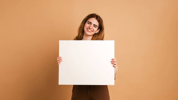 Portret van lachende jonge vrouw met donker haar met blanco plakkaat met mockup kijkend naar de camera terwijl ze tegen beige achtergrond staat - Foto, afbeelding