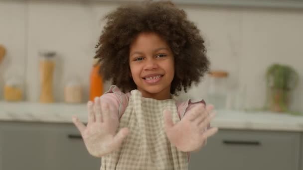 Αφρικάνικη αμερικανική λίγο χαρούμενος χαμογελώντας χαμόγελο θετικά παιχνιδιάρικο κοριτσάκι παιδί έθνικ παιδί στο σπίτι κουζίνα μαγείρεμα αρτοποιείο προετοιμάσει ζύμη αρτοσκευάσματα δείχνουν παλάμες πασαλειμμένο με λευκό αλεύρι - Πλάνα, βίντεο