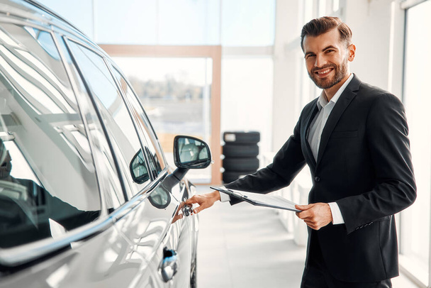 A salesman in a suit with documents stands in a car showroom and demonstrates the car to buyers, opening the doors. Buying, renting and insuring a car. - Photo, Image