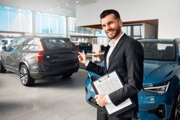 A salesman in a suit with forms of documents standing against the background of cars for sale in a car dealership. Purchase, rental and insurance of a car. - Photo, Image