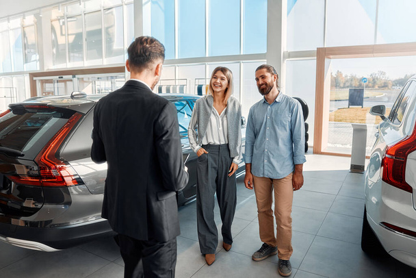 A salesman in a suit with a tablet consulting a couple of clients in a car dealership. Purchase, rental and insurance of a car. - Photo, Image