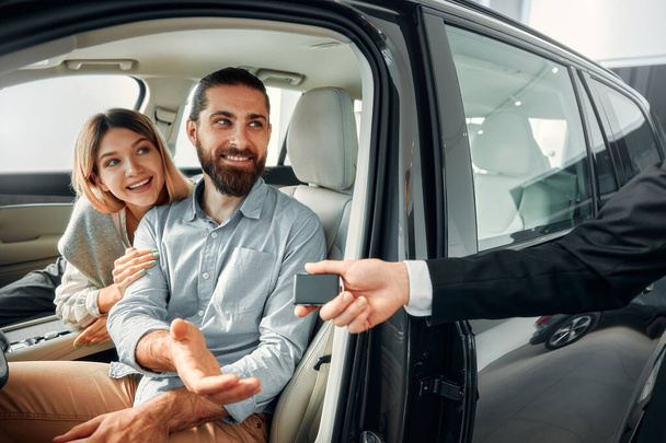 Šťastný mladý pár sedí v novém autě v autosalonu a dostává klíče od prodejce. Nákup, pronájem a pojištění vozu. - Fotografie, Obrázek