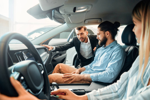 Una pareja está sentada en un coche en un concesionario de automóviles y recibiendo consejos de un vendedor. Compra, alquiler y seguro de un coche. - Foto, imagen
