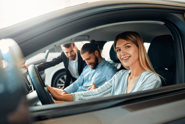 Una pareja está sentada en un coche en un concesionario de automóviles y recibiendo consejos de un vendedor. Compra, alquiler y seguro de un coche. - Foto, Imagen