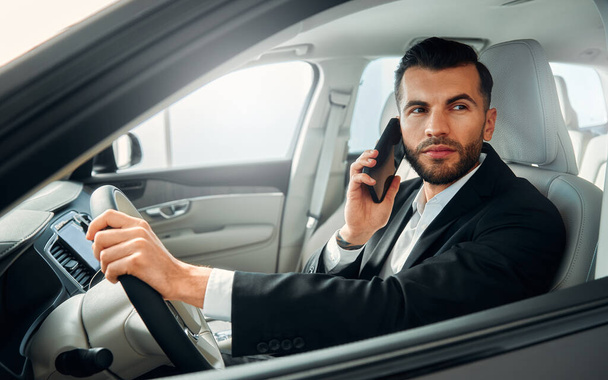 Άντρας επιχειρηματίας με μαύρο κοστούμι οδηγεί ένα αυτοκίνητο και μιλάει στο τηλέφωνο. - Φωτογραφία, εικόνα