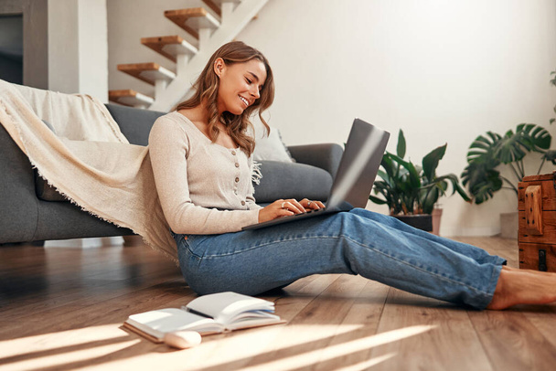 Mujer joven que trabaja en un ordenador portátil mientras se sienta en el suelo junto al sofá en la sala de estar en casa. Trabajo remoto y freelance. - Foto, imagen