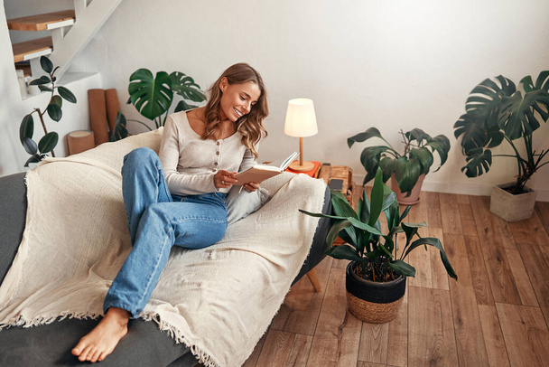 Młoda kobieta czyta książkę na kanapie w przytulnym salonie w domu, relaksując się i relaksując w wolny dzień. - Zdjęcie, obraz