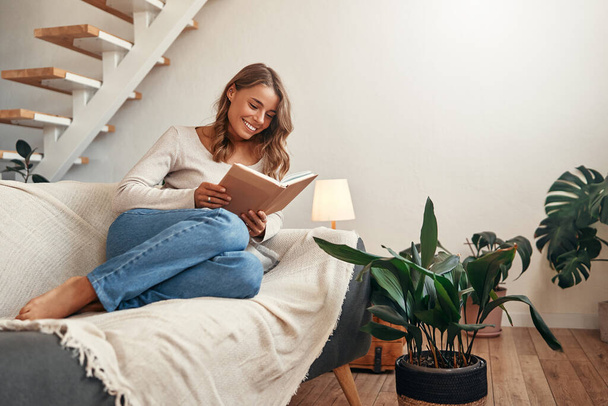 Junge Frau liest ein Buch auf dem Sofa in einem gemütlichen Wohnzimmer zu Hause entspannen und entspannen an ihrem freien Tag. - Foto, Bild