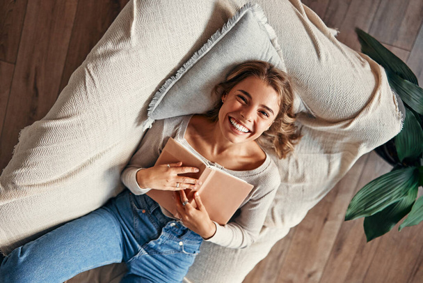 Молода жінка читає книгу на дивані в затишній вітальні вдома, розслабляючись і розмовляючи на вихідний. - Фото, зображення