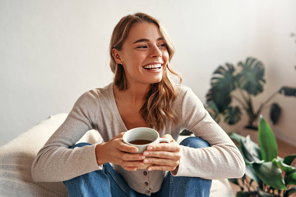 Fiatal nő egy csésze forró kávé vagy tea ül a kanapén egy hangulatos nappali otthon pihentető és lazító hétvégén nap. - Fotó, kép