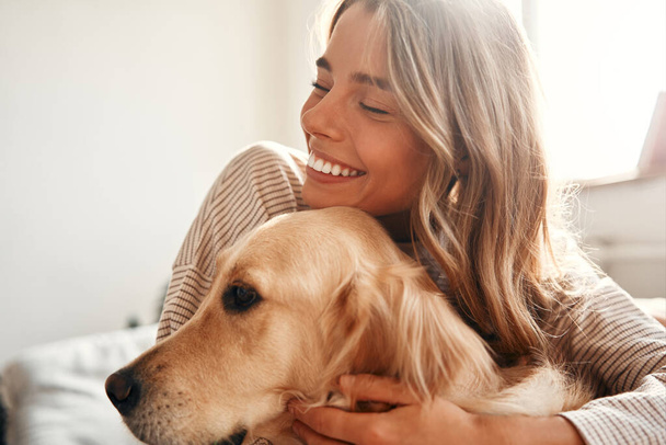 Ευτυχισμένη νεαρή γυναίκα με σκύλο στον καναπέ στο άνετο σαλόνι στο σπίτι χαλαρώνοντας και χαλαρώνοντας το Σαββατοκύριακο, αγκαλιάζοντας και παίζοντας με το κατοικίδιο ζώο της. - Φωτογραφία, εικόνα