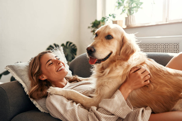 Mujer joven y feliz con perro en el sofá en la acogedora sala de estar en casa relajante y relajante el fin de semana, abrazando y jugando con su mascota. - Foto, Imagen