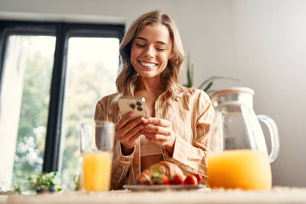 Jeune femme heureuse debout à la table dans la cuisine, à l'aide d'un smartphone, il y a des croissants avec des fraises dans une assiette, et un verre de jus. Femme prenant le petit déjeuner dans une cuisine confortable. - Photo, image