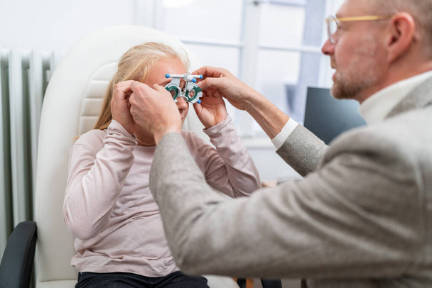 Optometrist fitting Optical measuring glasses on a young blonde girl during an eye exam at the ophthalmology clinic. - Photo, Image