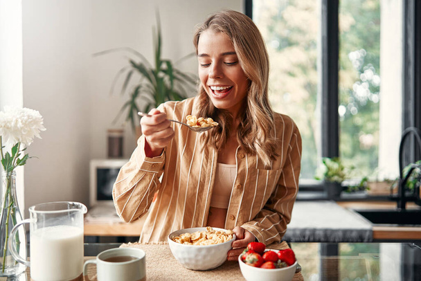 Young happy woman sitting at the table in the kitchen, eating cereal with milk, strawberries and drinking coffee. A woman is having breakfast in a cozy kitchen. - Photo, Image