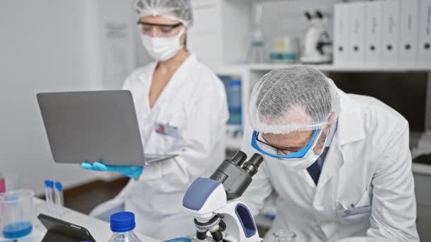 Juntos en el laboratorio, dos científicos luciendo máscaras médicas, acercándose con su microscopio mientras trabajan en su computadora portátil en medio de la investigación covid19. - Metraje, vídeo