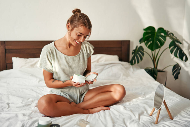 A young happy woman, waking up in the morning, sits on her bed, opening a jar of moisturizer. Skin care for face and body, morning routine, spa treatments. - Photo, Image