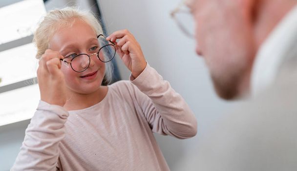 Jong meisje met een nieuwe bril en een glimlach. Opticien onderzoekt hoe een jong meisje glazen past in een brilwinkel.  - Foto, afbeelding