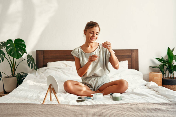 A young happy woman, waking up in the morning, sits on her bed, applying moisturizing serum to her face. Skin care for face and body, morning routine, spa treatments. - Photo, Image