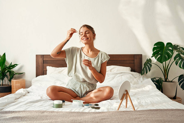 Eine junge, glückliche Frau, die morgens aufwacht, sitzt auf ihrem Bett und trägt ihrem Gesicht feuchtigkeitsspendendes Serum auf. Hautpflege für Gesicht und Körper, Morgenroutine, Wellness-Behandlungen. - Foto, Bild