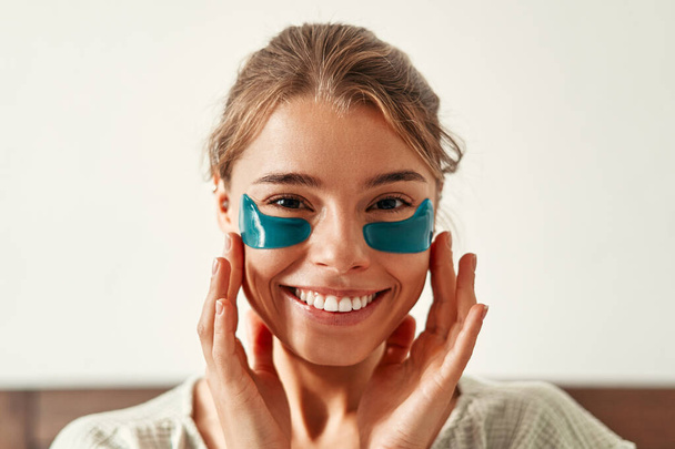 A young happy woman, waking up in the morning, sits on the bed with moisturizing patches under her eyes. Skin care for face and body, morning routine, spa treatments. - Valokuva, kuva