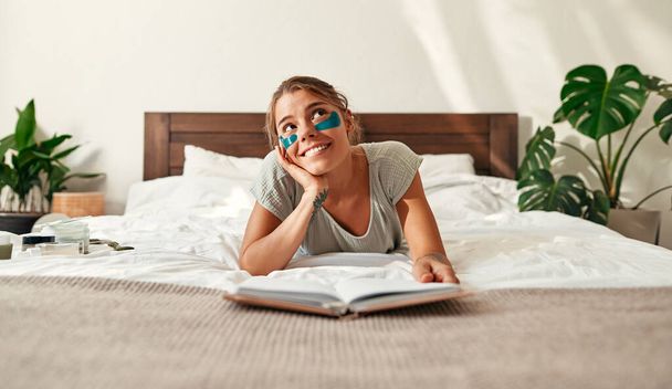 Nuori onnellinen nainen, joka herää aamulla, makaa sängyllä kosteuttavilla laastareilla silmien alla, lukemassa kirjaa. Kasvojen ja kehon ihonhoito, aamurutiini, kylpylähoidot. - Valokuva, kuva