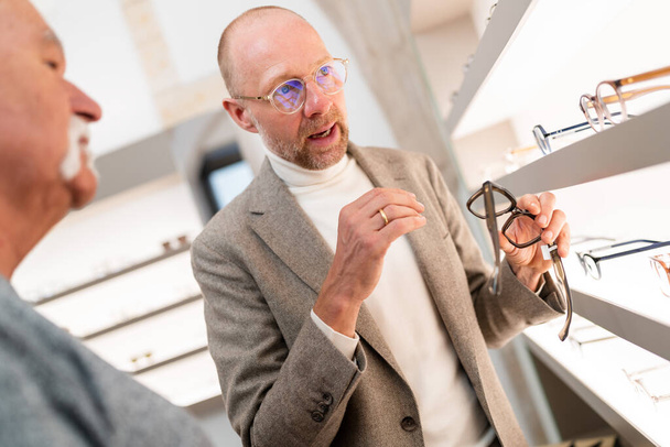 Optometrikus szemüveget mutat be az ügyfélnek az optikai boltban. Szemüvegpolc előtt mutatja be a szemüveg válogatást. - Fotó, kép