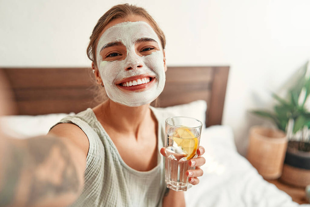 Een vrouw bracht 's morgens een klei gezichtsmasker aan en drinkt water met citroen terwijl ze thuis op het bed in de slaapkamer zit. Huidverzorging en spa-behandelingen. Handhaving van de waterbalans. - Foto, afbeelding
